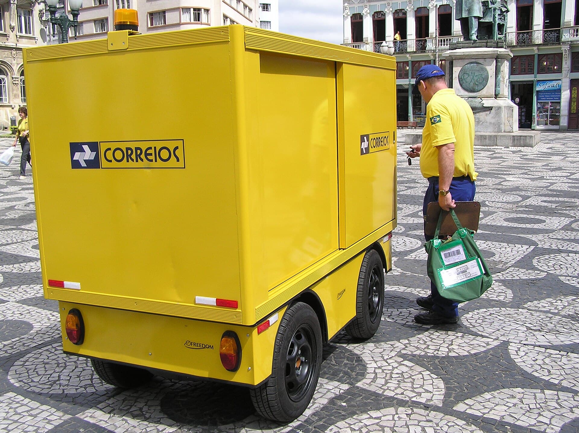 Carteiro dos Correios ao lado de um carrinho de entregas amarelo numa praça de calçada portuguesa